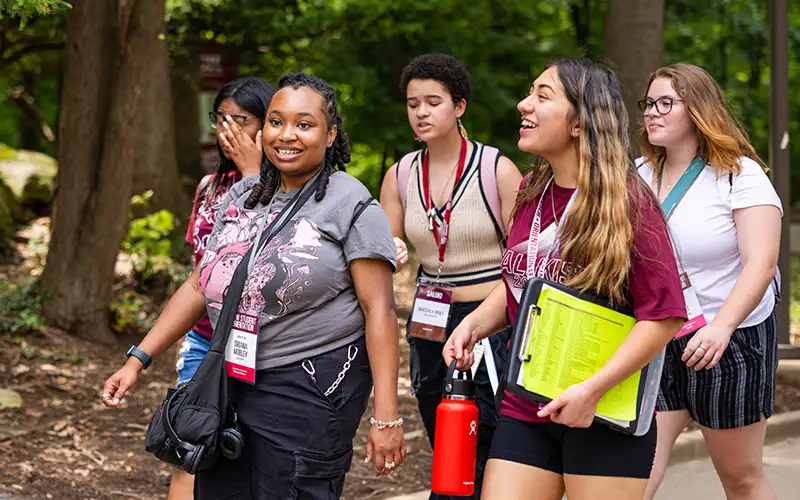 Group of students participating in a campus walking tour, smiling and chatting as they walk along a wooded path with lanyards and folders.