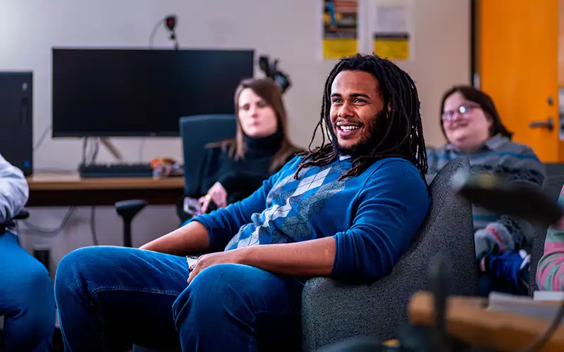 Student smiling while seated in a relaxed classroom or lab setting, surrounded by peers and computer workstations in the background.