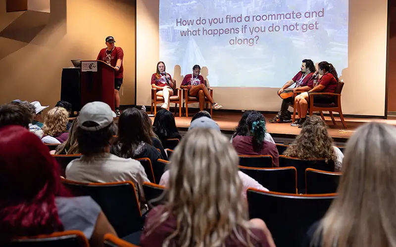 Students attend an information session in an auditorium where a panel discusses campus life, with a projected question about finding and getting along with roommates.