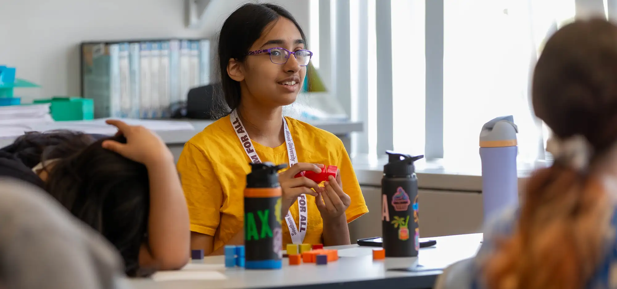 Student participating in an SIU field trip activity, seated at a classroom table with colorful blocks and water bottles, wearing a lanyard and speaking to peers.