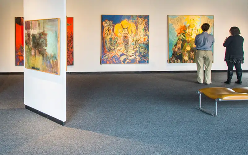 Two visitors view colorful abstract paintings on display in the Sharp Museum gallery, with art pieces mounted on white walls and a bench in the foreground.