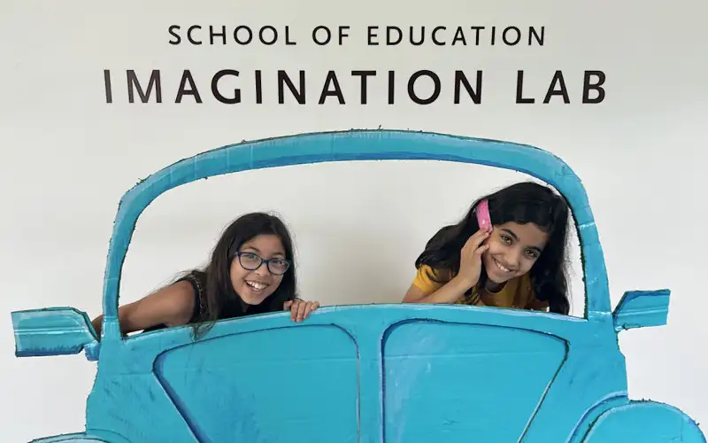 Two girls pose playfully behind a cardboard cutout of a blue car at the School of Education Imagination Lab, smiling and pretending to drive and talk on the phone.